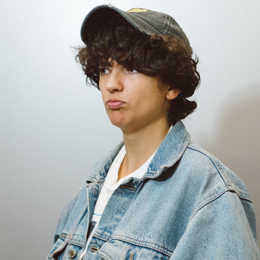 Retrato de perfil de la cantante Bari Ricanati, lleva una gorra gris, cabello corto y chino a la altura de las orejas y una camisa blanca con una chamarra de mezclilla azul claro, el fondo es liso y es gris