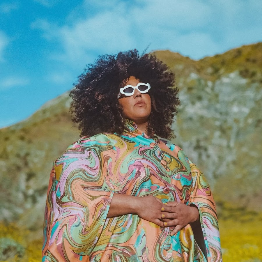 Una foto de la cantante Brittany Howard. Ella viste un vestido de color naranja, azul y verde, unos lentes de sol con armazón blanco, el cabello suelto y crespo y el fondo del paisaje es una montaña verde y un cielo azul con nubes